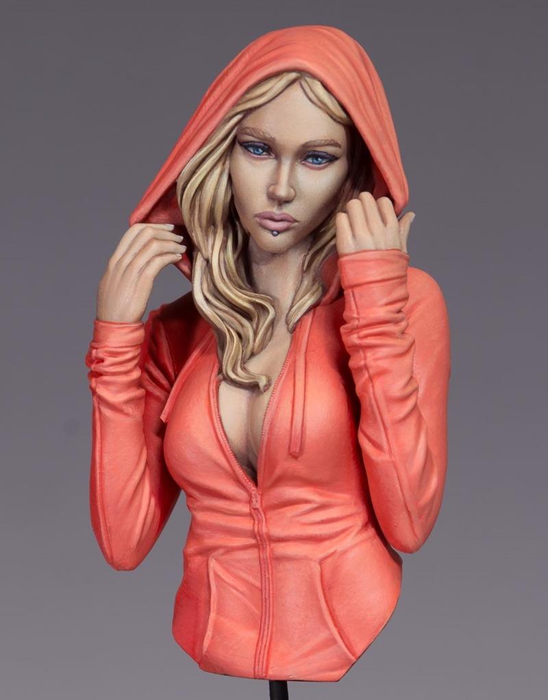 Bust of a blonde woman with blue eyes and a lip piercing, wearing an unzipped coral hoodie and adjusting the hood.