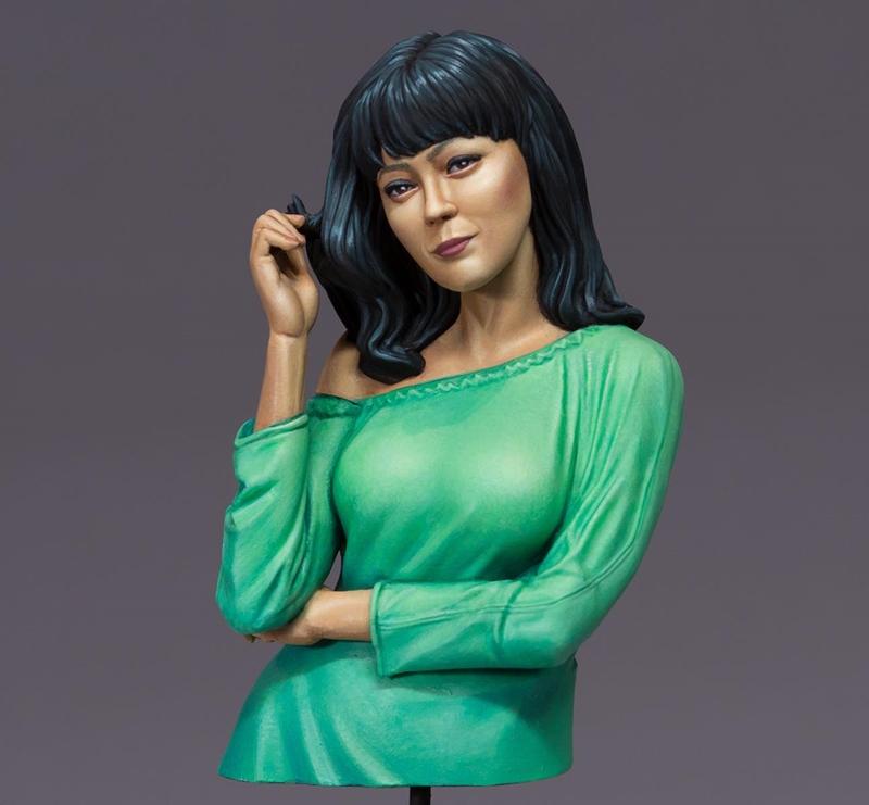 Bust of a woman with dark hair, wearing a green off-the-shoulder top, with one hand touching her hair and the other c...
