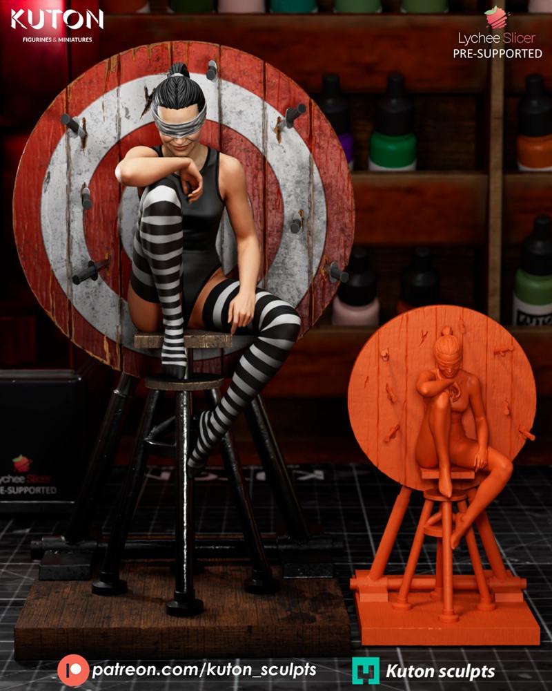 Blindfolded female figure 'Blind Trust' by Kuton Figurines, seated on a stool before a wooden target with knife detai...