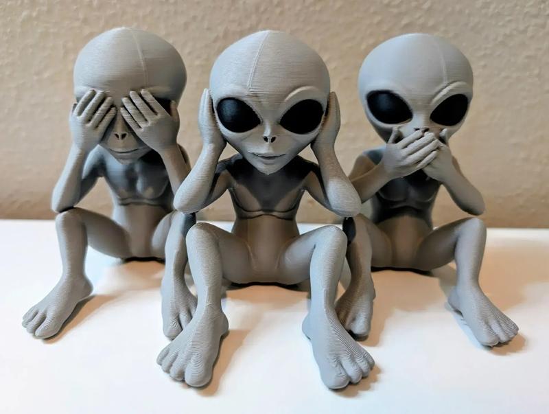 Three grey alien figures in a seated pose, representing 'see no evil' (covering eyes), 'hear no evil' (covering ears)...